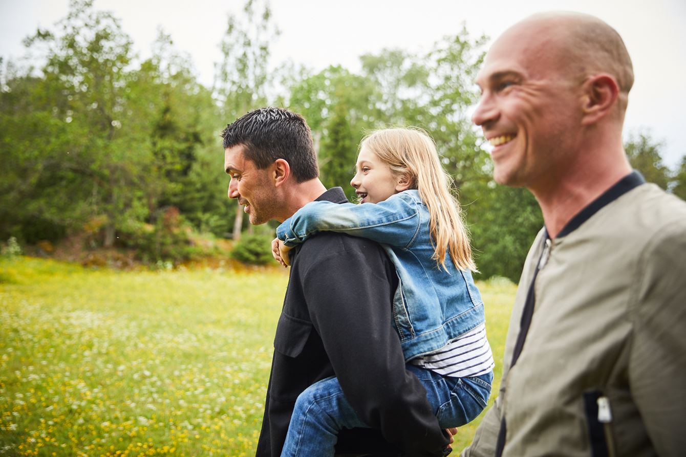 Två män på en äng, den ena mannen bär en flicka på ryggen